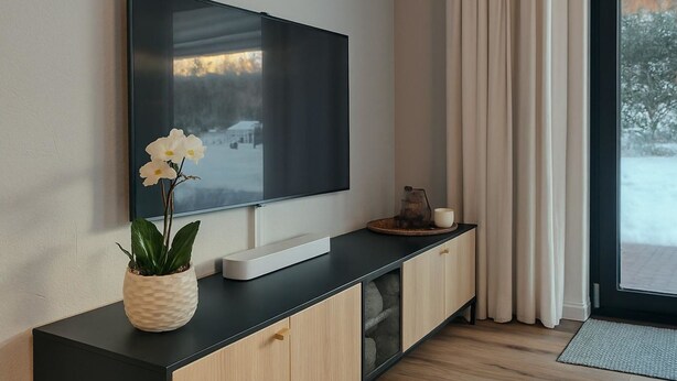 Un soggiorno moderno con una televisione a schermo piatto montata a parete, un mobiletto in legno con anta in vetro, un'orchidea in vaso e un tavolino con una tazza e un oggetto decorativo.