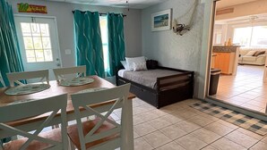 Ein Zimmer mit Bett, Deckenventilator, Esstisch mit Stühlen, Küchenzeile und Blick nach draußen.