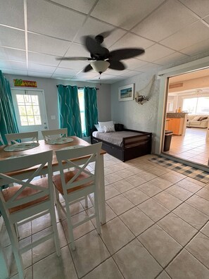 A room with a bed, a ceiling fan, a dining table with chairs, a kitchen area, and a view of the outside.