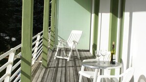 Um deck de madeira com cadeiras e mesa brancas, guarda-corpos verdes e um edifício branco com detalhes verdes.