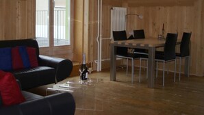 Um quarto com piso de madeira, um sofá de couro preto com almofadas vermelhas e azuis, uma mesa de jantar com quatro cadeiras e um radiador.
