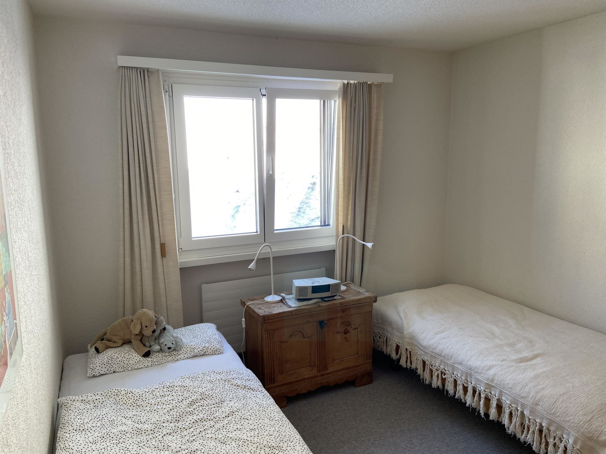 Ein Schlafzimmer mit zwei Betten, einer Holzkommode, einem Fenster mit Vorhängen und einer Heizung.