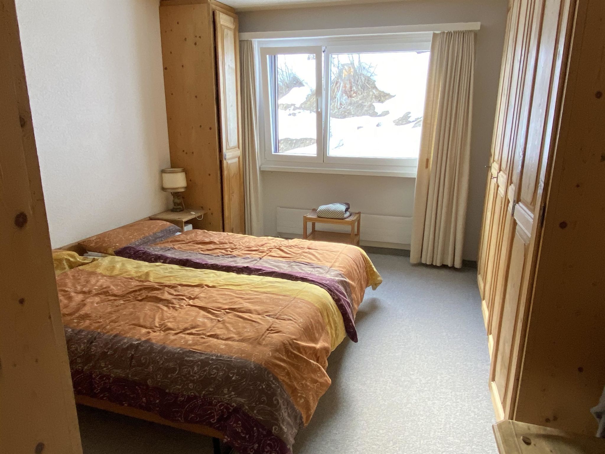 Un dormitorio con dos camas, una ventana con cortinas, una mesita de noche con una lámpara y una pequeña mesa con un cojín.