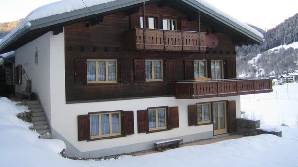 Un chalet en bois à deux étages, doté d’un balcon, de grandes fenêtres et d’un toit recouvert de neige.