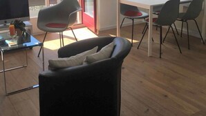 En moderne stue med sofa, spisebord med stole, fjernsyn og store vinduer med udsigt til træer.