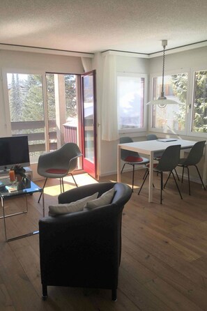 Ein modernes Wohnzimmer mit einer Couch, einem Esstisch mit Stühlen, einem Fernseher und großen Fenstern mit Blick auf Bäume.