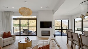 A spacious living room with a large sofa, a coffee table, and a fireplace. There are large windows offering a view of the mountains.