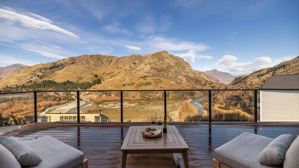 Terraza de madera con asientos exteriores y una mesa dispuesta con comida y bebidas, con vistas a un paisaje montañoso y un río.
