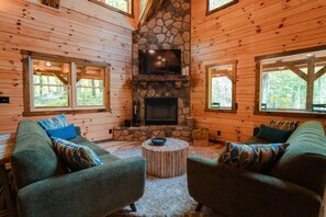 L’intérieur chaleureux d’un chalet avec des murs en bois, une cheminée en pierre, un téléviseur à écran plat et deux fauteuils verts.
