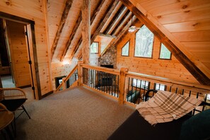 A wooden cabin interior with a stone fireplace, a balcony with a railing, and a view of the surrounding forest.