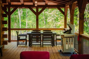 A covered outdoor area with a wooden table and chairs, a grill, and a view of a forest.