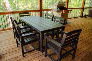 Um deck de madeira com mesa, cadeiras, churrasqueira e vista para árvores.
