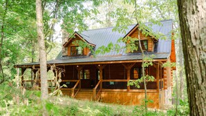 A log cabin with a porch, multiple windows, and a metal roof, surrounded by dense forest. - Knotty Summit| Hocking Hills, Theater, Hot Tub (Logan)