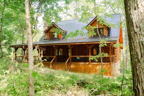 A log cabin with a porch, multiple windows, and a metal roof, surrounded by dense forest.