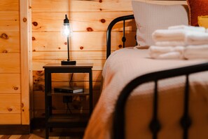 A wooden-paneled room with a bed, a nightstand with a lamp, and a shelf with books.