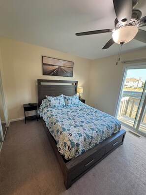 Un dormitorio con cama, ventilador de techo, mesita de noche, lámpara, un cuadro en la pared y vistas al exterior.