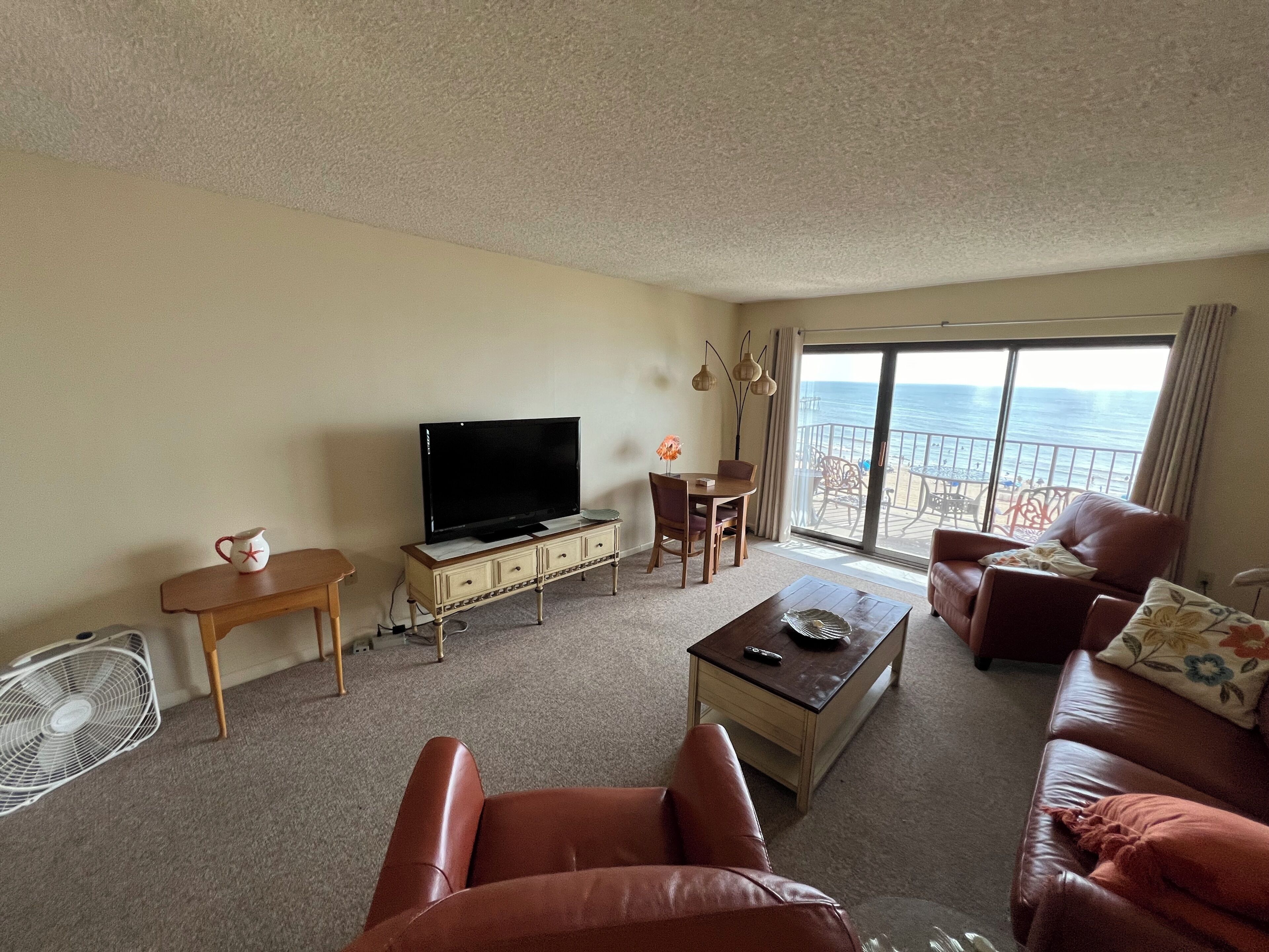 A living room with a TV, a dining table, a sofa, and a view of the ocean.