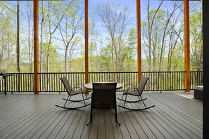 A wooden deck with a table and chairs, surrounded by a black railing, overlooking a forest.
