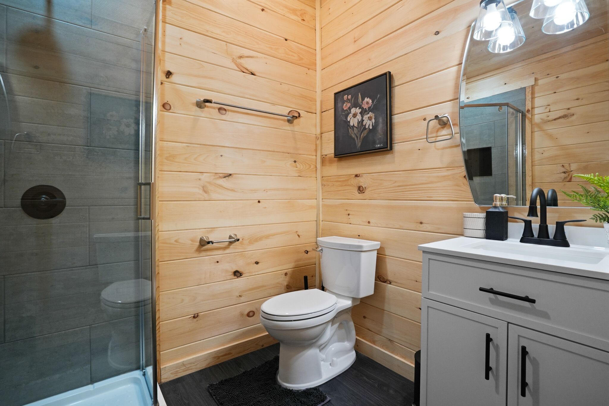 A wooden-paneled bathroom with a glass shower enclosure, a white toilet, a sink with a mirror, and a framed picture on the wall.