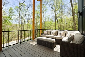 A covered outdoor seating area with a wicker sofa and ottoman, a black metal railing, and a view of a forested area.