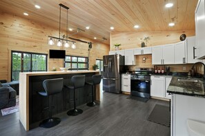 A modern kitchen with wooden walls, a central island with bar stools, stainless steel appliances, and a large window overlooking greenery.