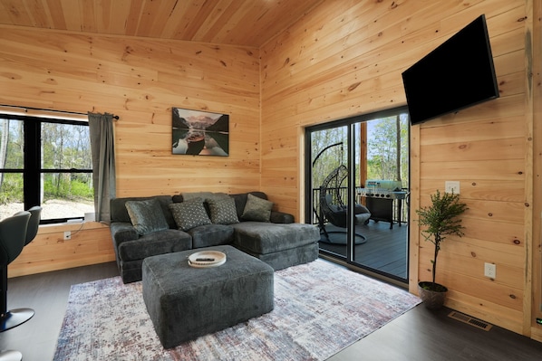A modern log cabin interior with a sectional sofa, a flat-screen TV, and a dining area visible through glass doors.