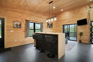 A wooden cabin interior with a bar area, three stools, a television mounted on the wall, and a large window.