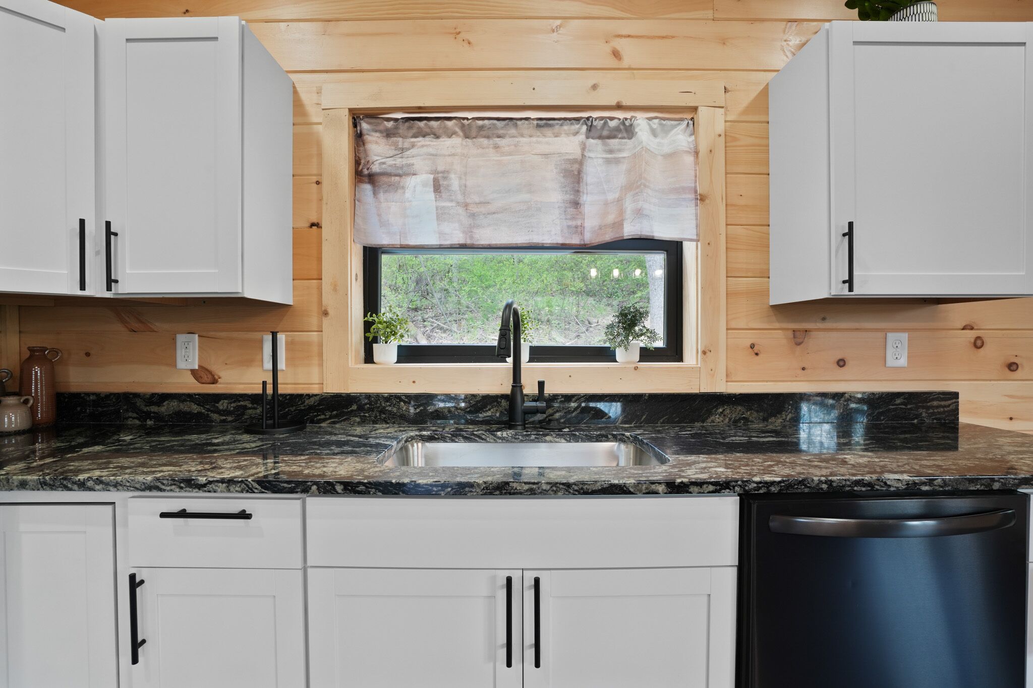 A kitchen with white cabinets, a granite countertop, a black faucet, and a window with a view of greenery.