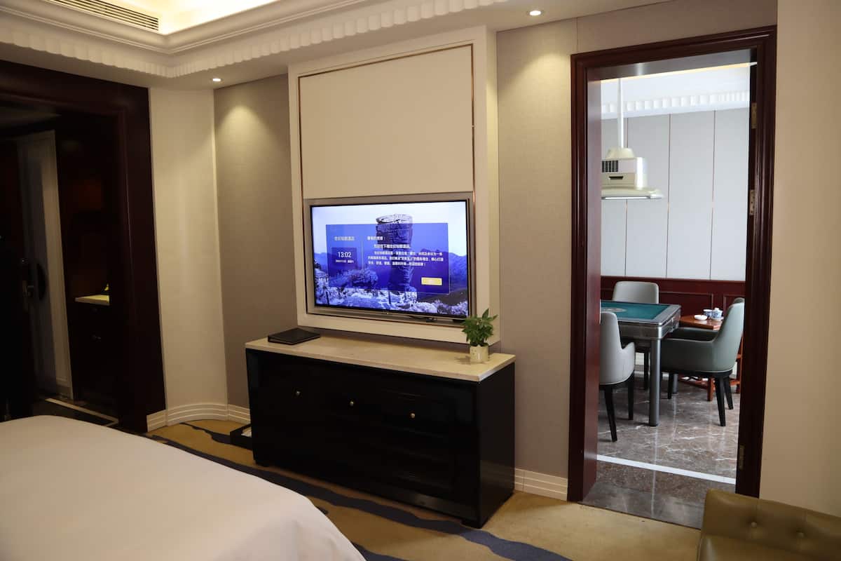 A hotel room with a bed, a TV mounted on a wall, a dark-colored console table, and a view of a dining area through an open door.