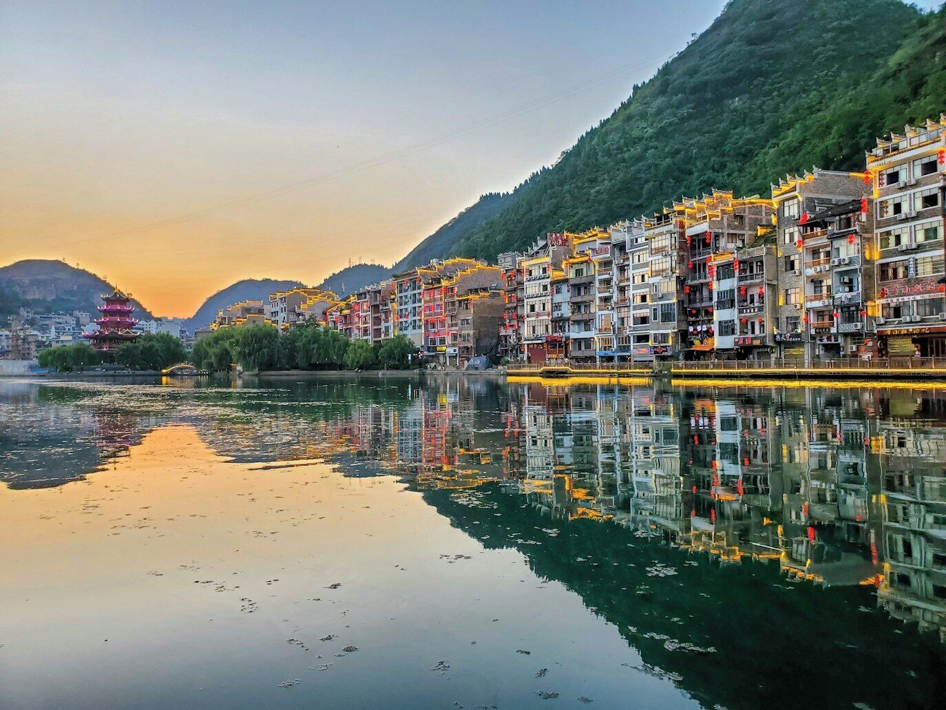 A riverside town with traditional architecture, a reflection in the water, and a mountain backdrop.