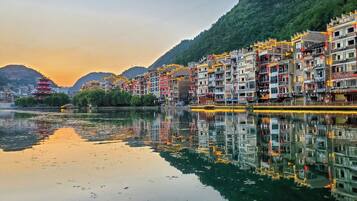 A riverside town with traditional architecture, a reflection in the water, and a mountain backdrop.