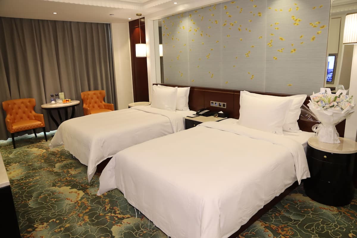 A hotel room with two beds, a small table with a chair, a nightstand, and a decorative wall.
