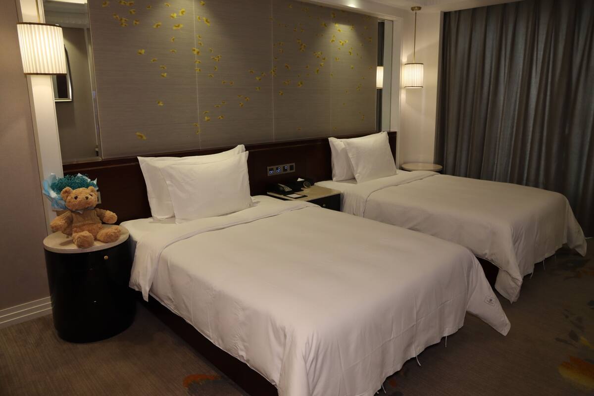 A hotel room with two beds, a nightstand, a teddy bear, and a wall with decorative elements.