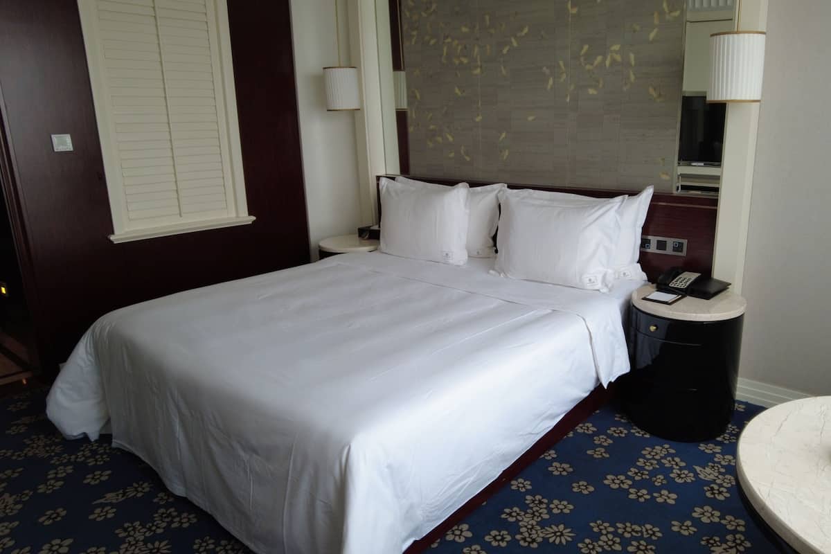 A hotel room with a large bed, a bedside table with a phone and a notepad, a round table with fruit, and a window with blinds.