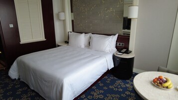 Quarto de hotel com uma cama grande, uma mesa de cabeceira com telefone e bloco de notas, uma mesa redonda com frutas e uma janela com persianas.