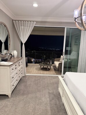 A bedroom with a balcony, a bed, a dresser, and a view of the city at night.