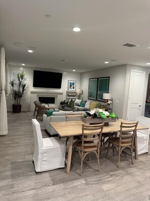 A modern living room with a flat-screen TV, a fireplace, a dining area with a wooden table and chairs, and a large window.