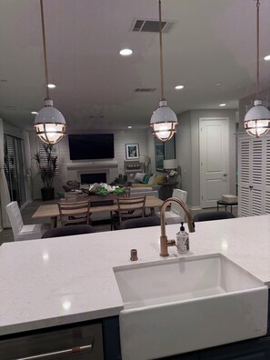 A modern kitchen with a white countertop, a stainless steel sink, and a copper faucet. There are pendant lights above the island, a dining area with wooden chairs and a table, and a TV mounted on the wall.
