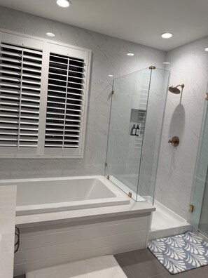 A modern bathroom with a glass-enclosed shower, a white bathtub, and a window with blinds.