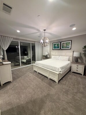 A bedroom with a large bed, a ceiling fan, a nightstand, a lamp, and a sliding glass door leading to a balcony.