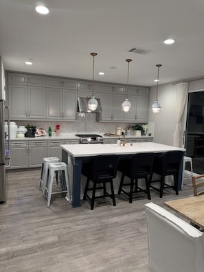 A modern kitchen with grey cabinets, a central island with bar stools, pendant lighting, and a stainless steel range hood.