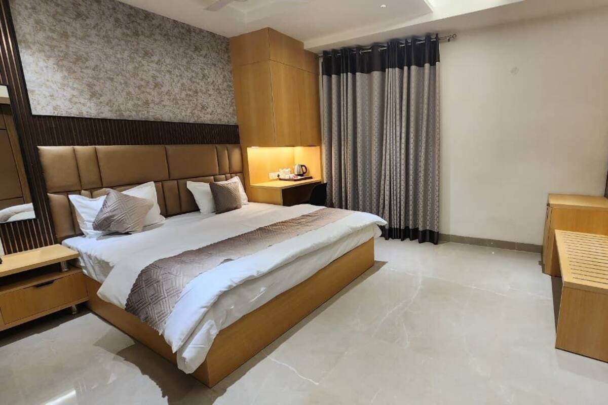 A modern hotel room with a large bed, wooden headboard, a bedside table with a lamp, a TV mounted on the wall, and a view of the cityscape.