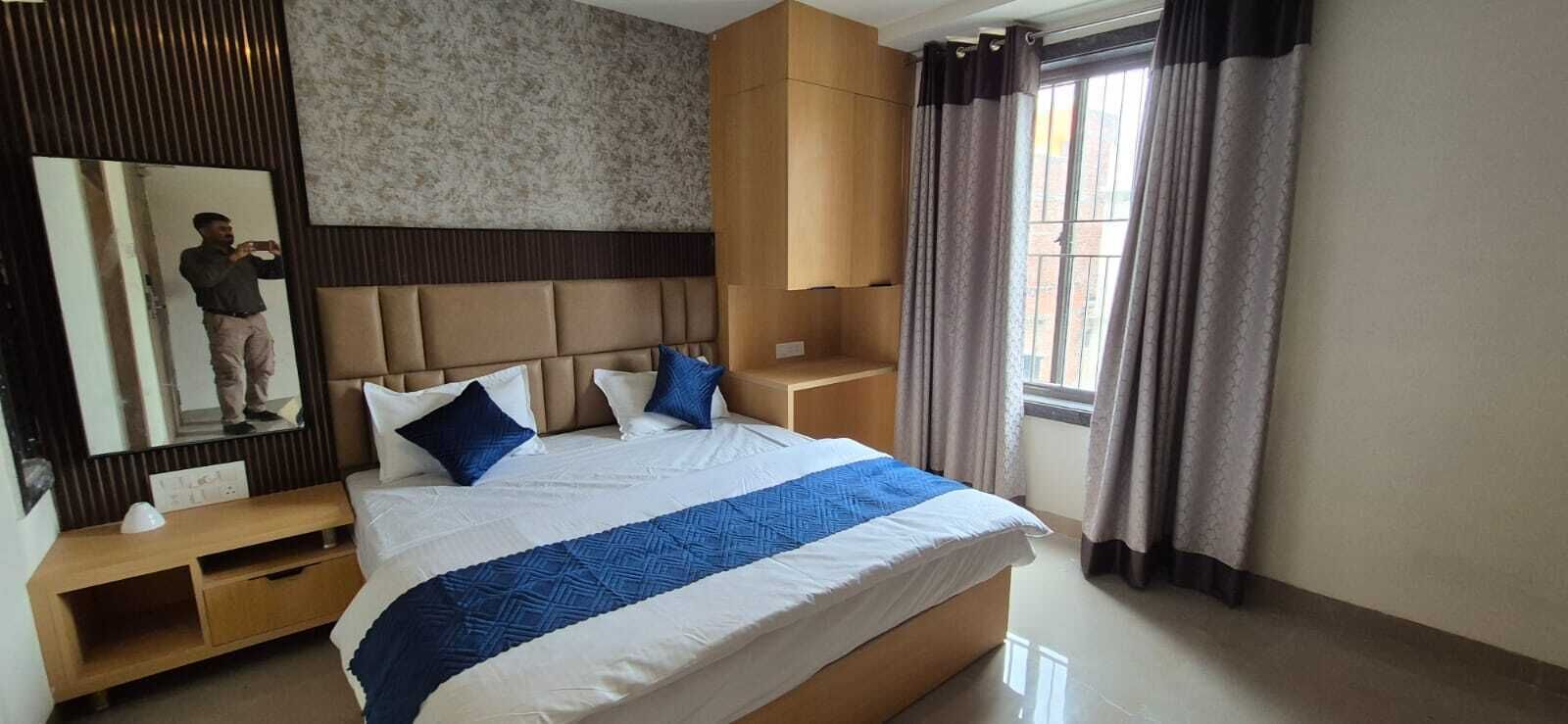 Une chambre d’hôtel comprenant un lit, un miroir, une table de chevet en bois, une tête de lit intégrée et une fenêtre avec des rideaux.