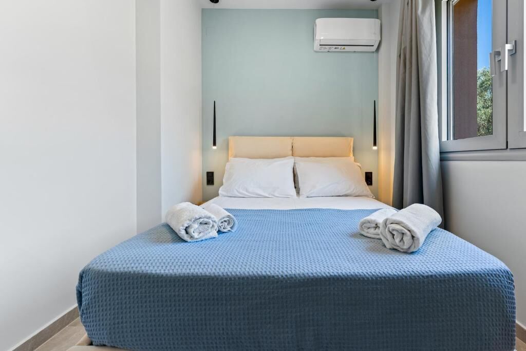 Une chambre d’hôtel avec un lit, deux oreillers, une couvre-lit bleue, deux serviettes enroulées, un climatiseur fixé au mur et une fenêtre avec des rideaux.