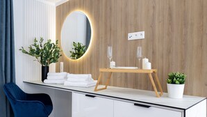A modern bathroom with a wooden vanity, a round mirror with lighting, a small table with glasses and candles, a blue chair, and a potted plant. - Studios an der Alten Post (Maxhütte-Haidhof)