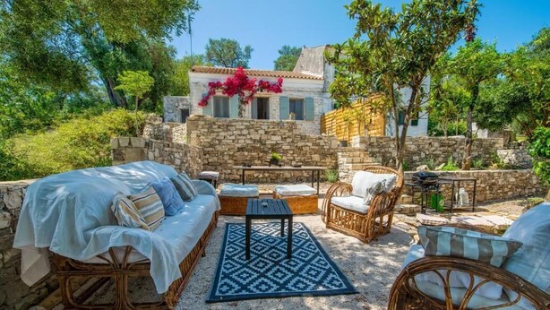 A stone-walled outdoor seating area with wicker furniture, a patterned rug, and a view of a building with shutters. - Malethonas Cottage by All About Paxos (Paxos)