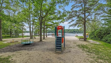 A playground with a swing set and a slide, surrounded by trees and a lake view.