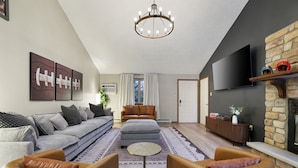 A modern living room with a sofa, ottoman, and leather chairs, a chandelier, a flat-screen TV, and a stone fireplace.