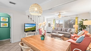 A living room with a wooden dining table, a television, and a sofa. - 🌊🌴Geaux Coastal | Charming 2BR Home | Steps to Private Beach Access (Navarre)