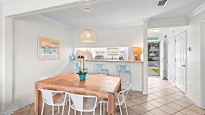 A dining area with a wooden table and white chairs, a hanging light fixture, and a kitchen in the background. - 🌊🌴Geaux Coastal | Charming 2BR Home | Steps to Private Beach Access (Navarre)